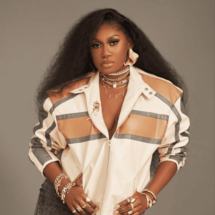 1000-x-1000-2025-05-02T161502.351.png Channeling Niniola’s vibrant style, a woman with long, textured hair wears a beige and brown striped jacket, layered necklaces, bracelets, and large earrings, standing confidently against a plain background.