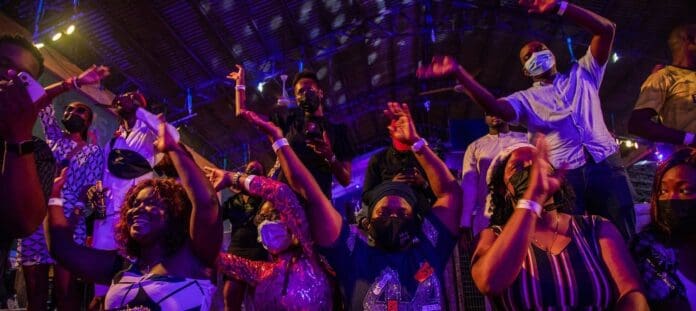 1500x6702Ffilters3Aquality2885292Fhttps3A2F2Fmedia.globalcitizen.org2Ff02Fa02Ff0a03051-53.jpeg A group of creatives wearing face masks cheer and raise their hands at an indoor event under colorful stage lighting in Lagos, Africa’s cultural capital.