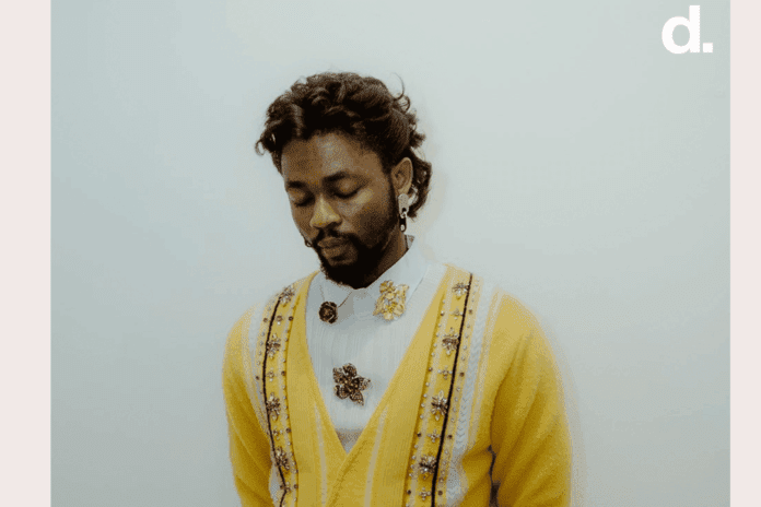 1d7fc54e-b493-48bc-8563-2c5e33f34875.png A man with dark curly hair and a beard looks down, wearing a yellow cardigan over a white shirt adorned with decorative brooches, channeling Afrobeats vibes like Omah Lay, against a plain light background.