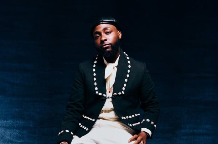A man wearing a black studded jacket, white shirt, and black leather cap sits against a dark background, looking at the camera with a neutral expression—much like an artist ready for his Billboard debut on the U.S. Afrobeats Songs Chart.