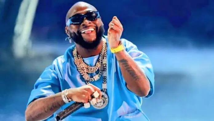Davido-1424x802-1.jpg A man wearing sunglasses, blue clothing, and multiple chains smiles while holding a microphone on stage, embodying the vibrant energy of Afrobeats.