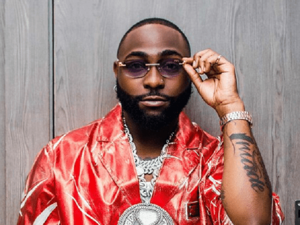 Davido.png A man with a trimmed beard wears a shiny red patterned jacket, sunglasses, and silver jewelry, posing in front of a wooden background—a style often seen in UK media and Afrobeats-inspired West African music scenes.