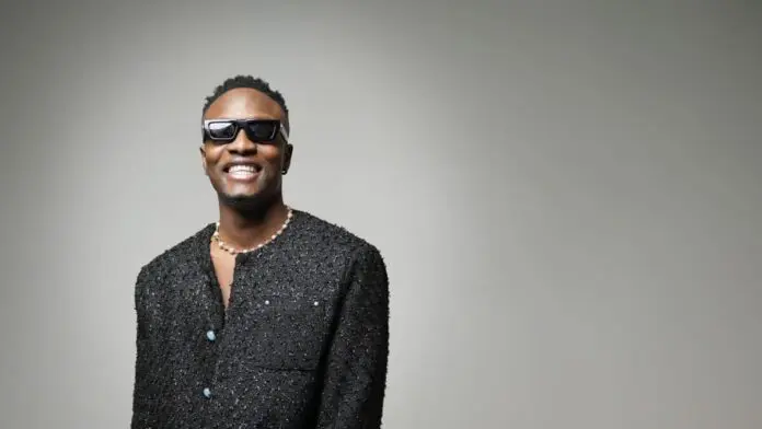 Driz-1.jpeg A person wearing dark sunglasses and a textured black jacket stands against a plain gray background, smiling at the camera with the confident energy of a Nigerian Afrobeats star.