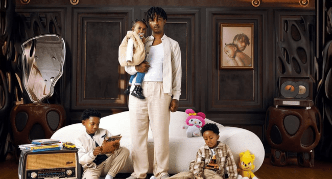 Emtee-14.png A man stands in front of a couch holding his daughter Nairobi, with two children sitting on the floor playing with devices; framed portraits and plush toys decorate the room, creating a perfect scene for a heartfelt birthday tribute.