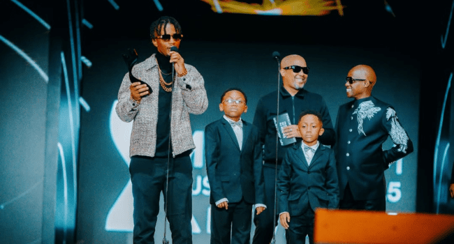 Emtee-15.png A group of five people, including three children and two adults in suits, stand on stage at the Metro FM Awards. One adult, inspired by Emtee’s Twice the Pressure album, holds a trophy and speaks into a microphone.