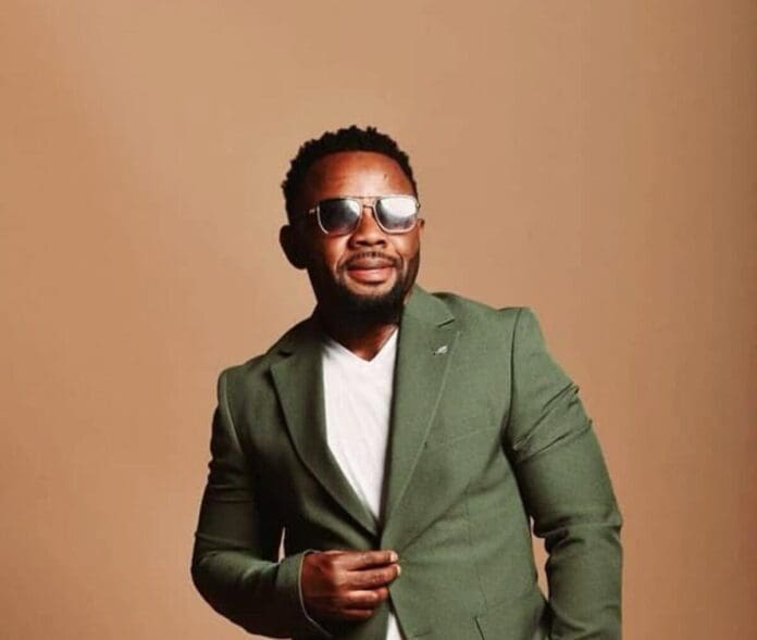 Expose-Talent-Records-1-e1746273613463.jpg A man wearing sunglasses, a green blazer, and a white T-shirt stands against a plain beige background, posing with one hand adjusting his blazer—exuding the confident style often seen in the African music scene.
