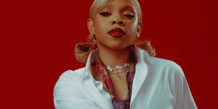 Fave-1440x720.jpg Person with blonde hair styled in pigtails, wearing a white shirt, red lipstick, and layered necklaces, posing against a solid red background—ready to channel the vibe of your fave hit tracks.