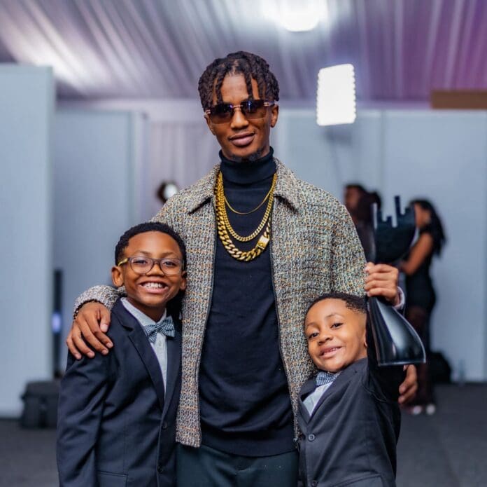 IMG_0744.jpeg A man wearing sunglasses and gold chains stands indoors with his arms around two smiling boys in suits; one boy proudly holds up a 2025 Metro FM Music Awards trophy.