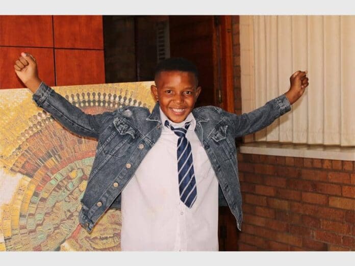 IMG_2464_76720.jpg A smiling boy in a denim jacket and striped tie stands indoors with his arms raised, channeling the vibrant energy of amapiano, in front of a colorful painting and brick wall.