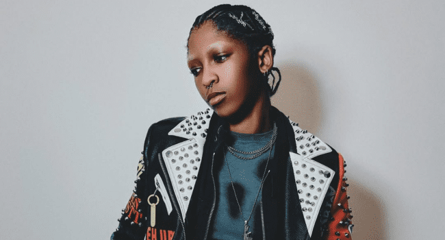 K.Keed-11.png Person with braided hair wearing a studded leather jacket and layered necklaces, looking to the side against a plain background, channeling K.Keed’s “Bite The Bullet” vibe for that milestone success attitude.