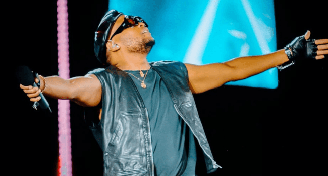 K.O-10.png A man wearing sunglasses, a leather vest, gloves, and a hat holds a microphone and stretches his arms out wide on stage under bright lights at the MMA25 King of the Culture event.