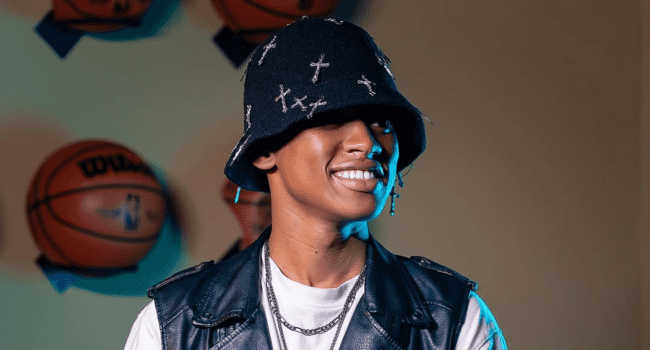 Kane-Keid-4.png A person wearing a black bucket hat with cross designs, a black leather vest, and a white shirt smiles with basketballs mounted on the wall in the background, showcasing Kane Keid’s Not Famous EP tracklist style.