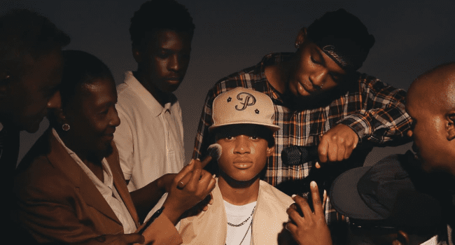 A young man in a beige hat and jacket, Kane Keid, is surrounded by people applying makeup, adjusting his clothing, and holding a microphone under dramatic lighting—capturing the behind-the-scenes energy of his Not Famous EP music shoot.