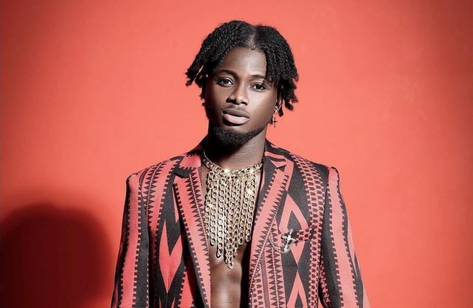 A man stands against a red background wearing a patterned jacket, layered chain necklaces, and sporting short twisted hair.