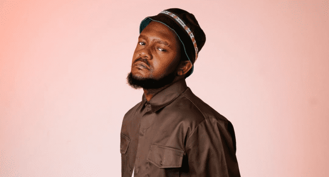 Kwesta-8.png A man with a beard, dressed in a brown button-up shirt and bucket hat, stands against a plain light pink background—echoing the laid-back vibe of Kwesta’s The Big Bro Theory upcoming album.
