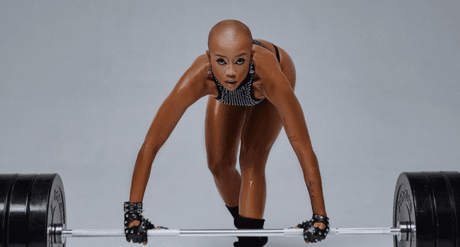 Moozlie-8.png A bald, Go Getter woman wearing gloves and workout attire prepares to lift a heavy barbell, positioned in a weightlifting stance on a plain background.