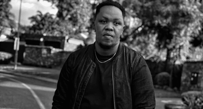 PdotO-8.png A man wearing a dark jacket and shirt stands outdoors on a street, with trees and a fence in the background. The black and white image has the cinematic feel of a PdotO music video.