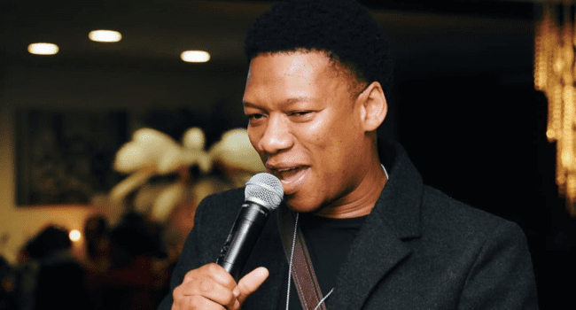 Proverb-3.png A man in a dark jacket, possibly ProVerb, speaks or sings into a microphone indoors, with blurred lights and decor in the background—capturing the vibe of a Prestigious Urban Music Award event.