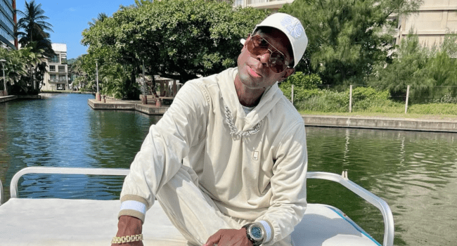 Rashid-Kay.png A man dressed in light-colored clothing and accessories sits on a boat by a canal, channeling the laid-back vibe of a classic hip-hop album, with trees and buildings in the background.