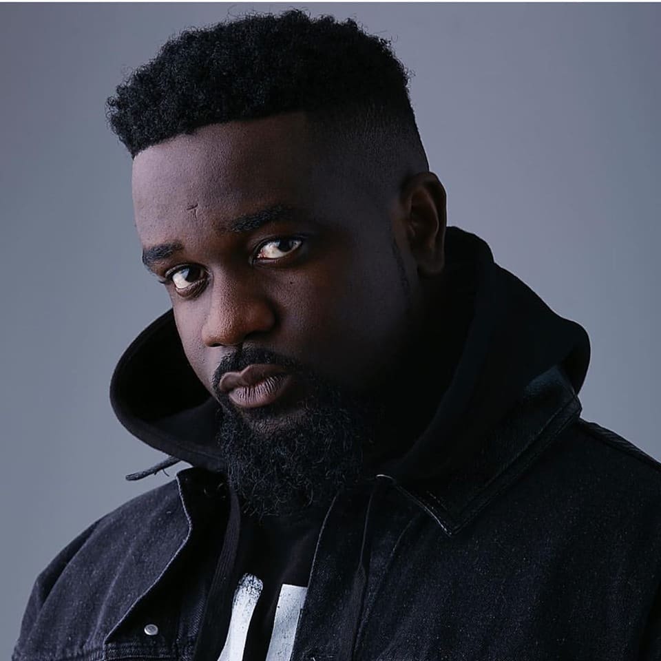 A man with short curly hair and a beard wearing a black hoodie and black denim jacket, looking directly at the camera against a plain gray background.