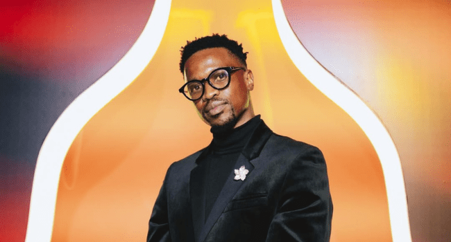Scoop.png Scoop Makhathini, wearing glasses and a black suit, stands in front of a brightly lit, orange and white abstract background as he opens up about fatherhood.
