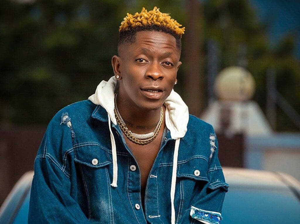 A man with short blond-tipped hair wears layered gold chains, a white hoodie, and a distressed denim jacket, standing outdoors with blurred background.