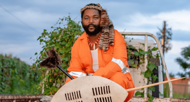 Sjava-9.png A man in an orange jumpsuit and traditional headpiece, perhaps a true Sjava fan, sits holding a shield and spear in an outdoor setting with greenery—echoing themes from the ATM Album.