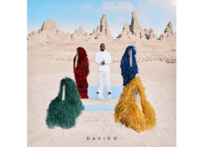 WhatsApp-Image-2025-05-04-at-14.30.41.jpeg A man in a smooth, stylish white suit stands in a desert landscape, surrounded by four figures in colorful, fringed costumes; rocky peaks rise behind them, echoing the bold visuals of Davido’s 5ive.