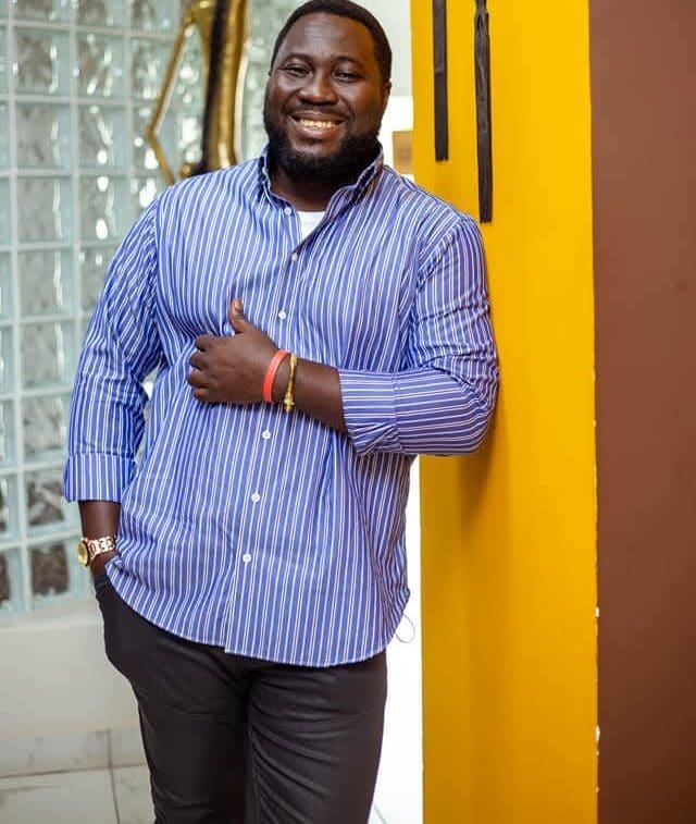 WhatsApp-Image-2025-05-05-at-1.09.43-AM-e1746444983637.jpeg A man in a blue and white striped shirt and dark pants stands indoors, leaning against a yellow wall, smiling with his thumb up—ready to enjoy some Afrobeats tunes on Asaase Radio.