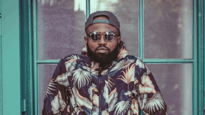 Wiziboy_20250503_095017_0000.png A man with a beard wearing a floral jacket, sunglasses, and a backward cap stands in front of a teal-framed window, channeling K1nxley's bold style from his new single "Recipe.