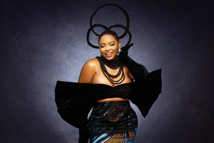 Yemi20Alade20002.jpeg A woman poses against a dark background, wearing a bold black top with large sleeves, layered bead necklaces, a patterned skirt, and braided hair styled in large circular shapes—her look echoing the fearless style of Palestinian Star Saint Levant.