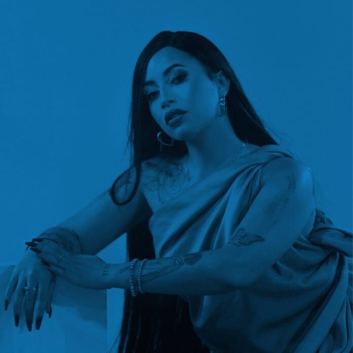 attachment-2-scaled.jpeg A woman with long dark hair and tattoos poses in a draped dress, resting her arm on a surface; the blue-tinted image captures a vibe reminiscent of Rowlene’s upcoming EP.