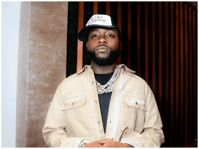 cd7c94ab-3c6d-4705-8313-f90f85ae4d5f.png A man wearing a beige jacket, black shirt, layered silver necklaces, and a white-and-black cap stands indoors in front of vertical wooden slats, resembling a scene from a Davido music video.