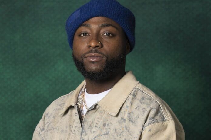 d71e22412af43b48f19ad92296e454364a445a35039e538c5c22a66f60502f28.jpg A man with a short beard and nose ring wears a blue beanie, beige jacket, and white shirt, posing against a textured green background with subtle Afrobeats vibes.