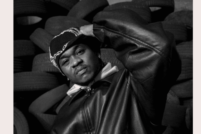 f2314db6-ee2a-4113-97d3-aa686ffa15ef.png A person wearing a beanie and leather jacket poses in front of a stack of tires, channeling Afropop vibes and looking directly at the camera with a neutral expression.