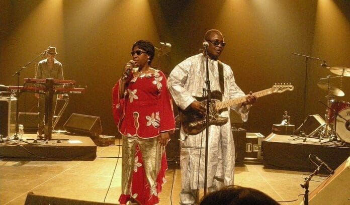 full.jpg Two musicians perform on stage; the woman sings into a microphone, and Malian musician Amadou Bagayoko plays electric guitar. Both wear traditional clothing. A keyboard player and drums are visible in the background, evoking West African music.