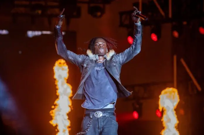 rema-live-aug-2024-billboard-1548.jpg A performer on stage raises both arms as flames shoot up in the background during Rema's electrifying Afrobeats live concert with dramatic lighting.