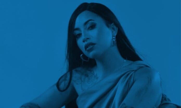 rowlene.jpeg Rowlene, with long dark hair and hoop earrings, poses in a draped outfit against a solid blue background, celebrating her new single.
