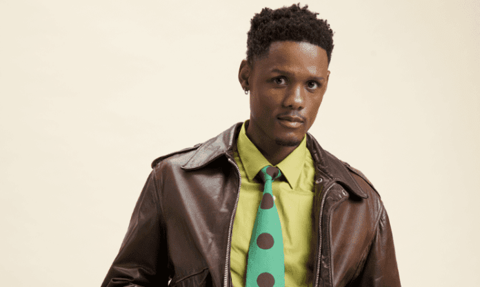 samthingsoweto1.png A person wearing a brown leather jacket over a lime green shirt and a matching green tie with large brown polka dots stands against a plain light background, ready for an a cappella show in Joburg.