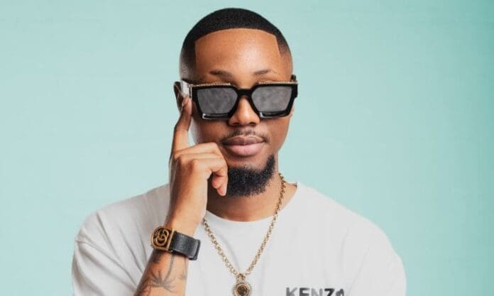 shaunstylist.jpeg Shaun Stylist, wearing dark sunglasses, a white shirt, a gold necklace, and a watch, poses against a light blue background, touching his sunglasses with one hand—hinting at the cool confidence ahead of his second EP.