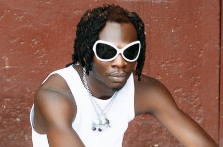 wp-17460091802045073780280696017492.jpg A person with braided hair wears large white sunglasses, a white tank top, and silver necklaces, striking a Body Magic pose in front of a textured reddish-brown wall.