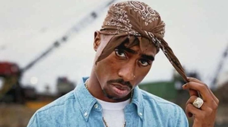 Tupac Shakur wearing a blue denim shirt and a brown bandana, channeling hip hop style, looks at the camera while adjusting his bandana, with an outdoor industrial background.