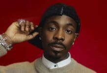 From Producer to Star: Young Jonn Defines the Sound of Modern Afrobeats – OkayAfrica A person wearing a beige sweater, white collared shirt, jewelry, and a black cap poses against a red background, channeling the vibrant style seen in OkayAfrica’s features on Modern Afrobeats artists like Young Jonn.