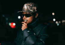 A-Reece Motions Music Video Shoot With LaCabra After Ignoring Stogie T’s “Four Horsemen” Call Person wearing sunglasses, a camouflage cap, and a dark puffer jacket standing outdoors at night with blurred city lights in the background, reminiscent of scenes from the Motions Music Video by A-Reece and Stogie T.