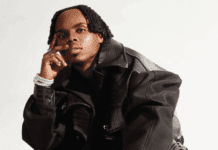 Ndu Browns Prepares To Release Solo Single “Summer Rain” A person wearing a black textured jacket poses seated against a plain white background, channeling the reflective vibe of Ndu Browns' solo single, with one hand resting on their face and the other on their knee.