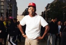 African Music Samples in Rap, R&B Songs: Missy Elliott, Rihanna & More – Billboard A man in a white T-shirt, red cap, and glasses stands outdoors with hands on hips, surrounded by people at an event featuring lively rap songs and African music samples.