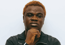 Diamond Boy Ignites Street Spirit on Raw Afro Streethop Anthem ‘Maji’ A person with short, curly, light brown hair wearing a black jacket poses against a plain white background, embodying the cool confidence of an Afro Streethop artist while resting their chin on their hand.