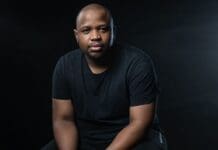 A beginner’s guide to breaking into the South African music industry – Music In Africa | A man wearing a black t-shirt sits against a dark background, looking directly at the camera with a neutral expression, as if ready to share insights from a beginner’s guide to the South African music industry.