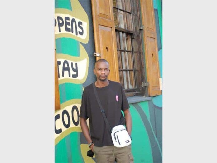 SFDGB_17837.jpg A music producer stands in front of a vibrant mural and a wooden window, wearing a black t-shirt and crossbody bag, his style echoing the rhythm of Amapiano beats.