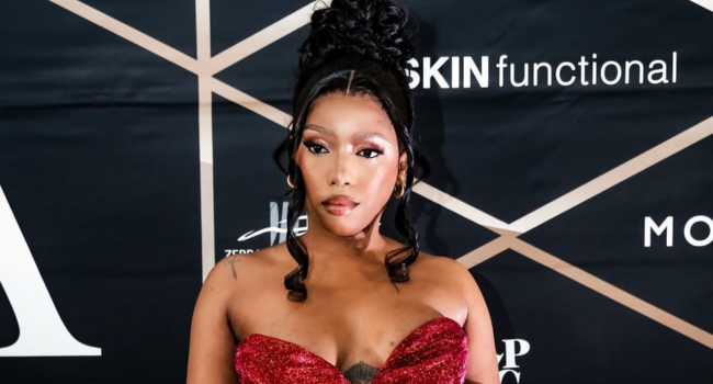 Gigi-Lamayne.png Gigi Lamayne, in a red strapless dress, poses confidently in front of a black backdrop with white geometric lines and various brand logos.